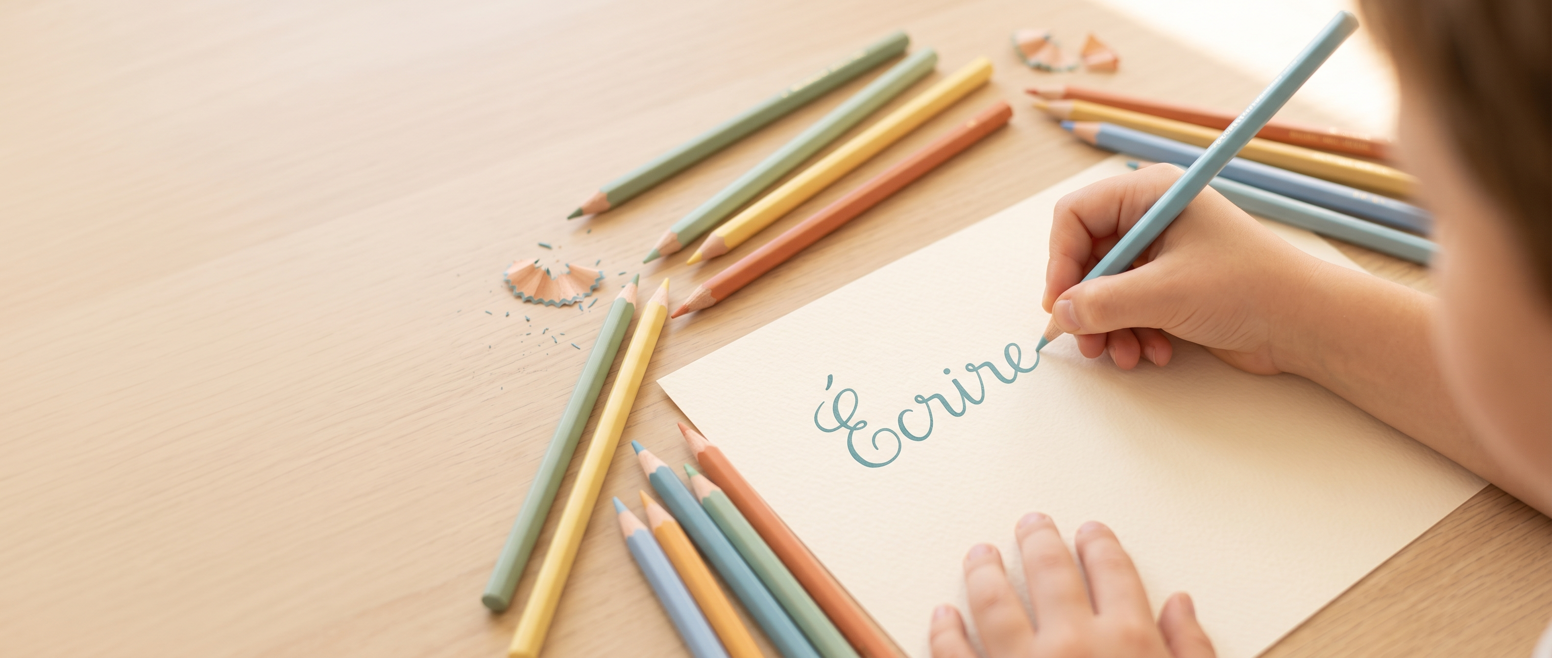 Main d'enfant écrivant le mot « Écrire » en lettres cursives, entourée de crayons de couleur aux tons pastel posés sur un bureau en bois clair.